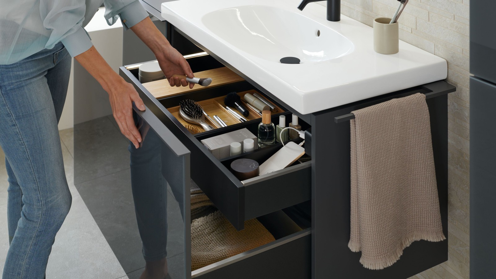 Woman reaches for a brush from an open Geberit Acanto washbasin cabinet in a lava finish